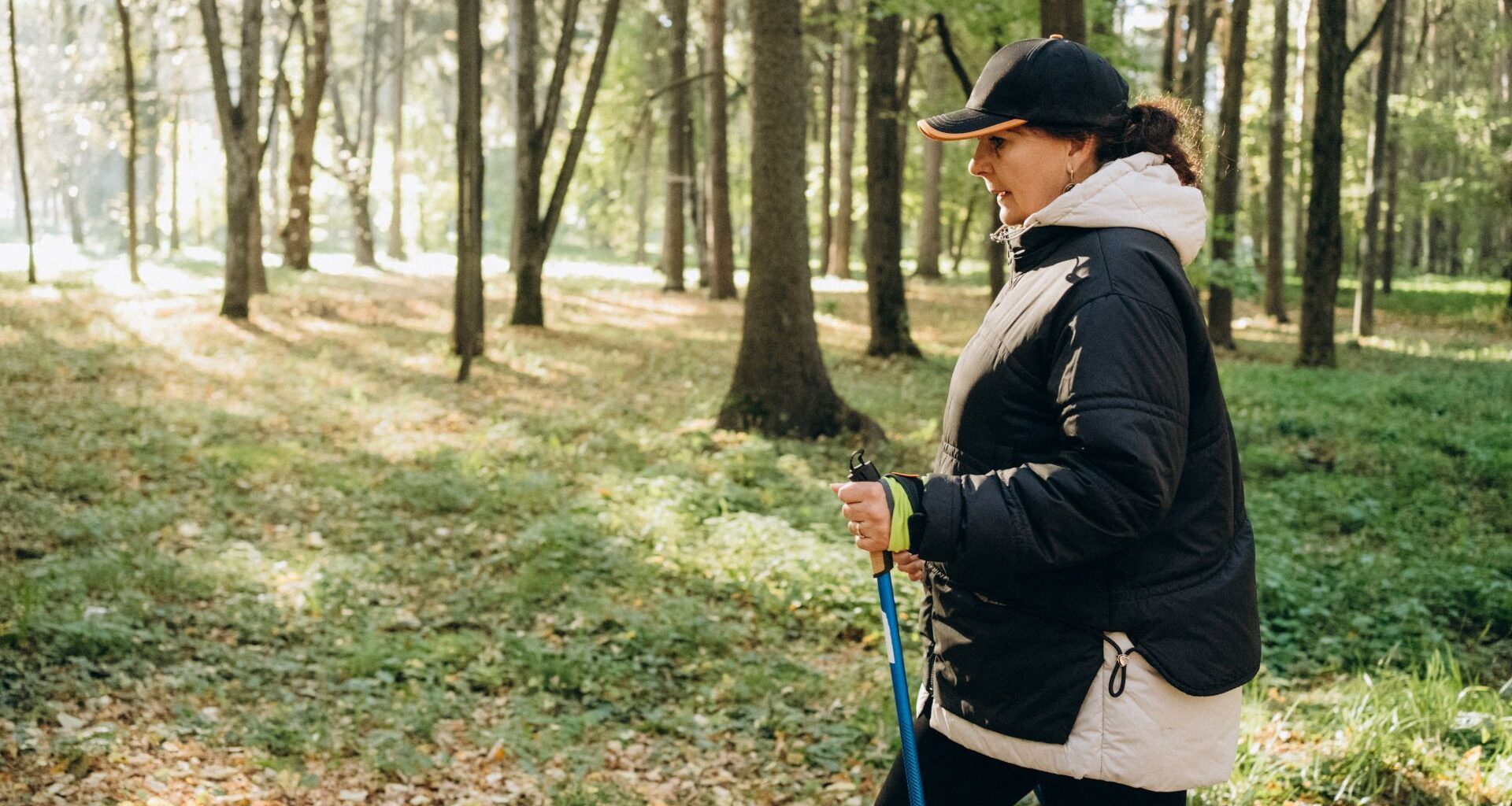 Woman doing metabolic walking holding pole and walking through forest wearing activewear and cap with coat on