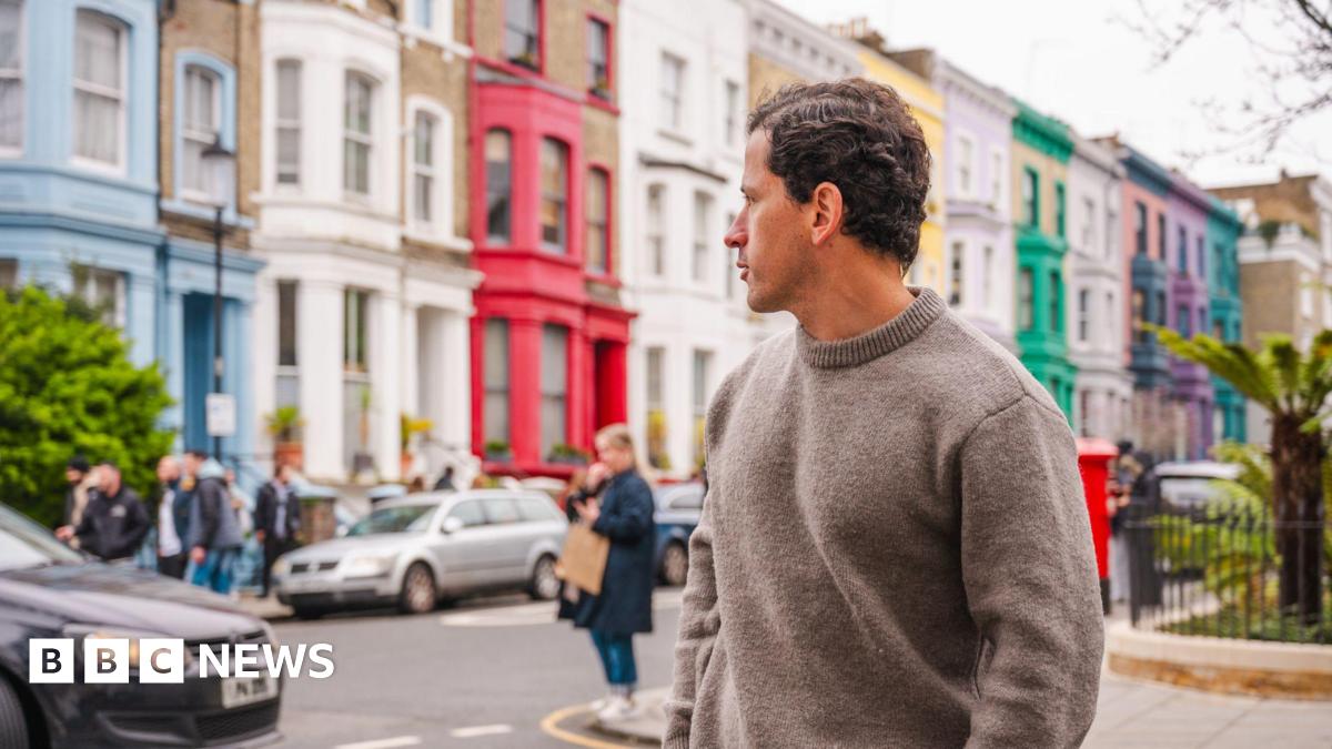 Young man in a jumper looks back over his shoulder at a row of brightly painted terraced Notting Hill townhouses.