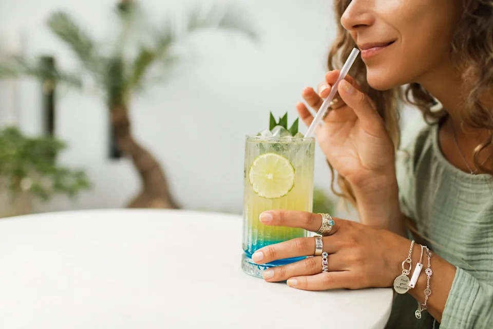 Woman sipping a layered cocktail with lime garnish through a straw at a white table, wearing rings and bracelets