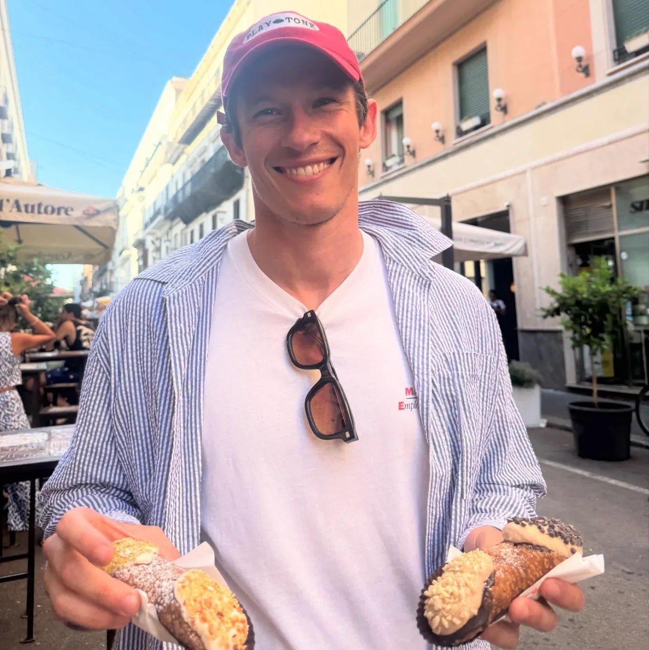 Man smiling and holding two cannolis on a street.