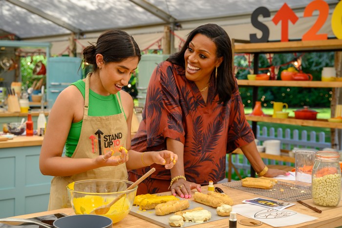 Ambika Mod smiling down at a work service whilst she bakes whilst Alison Hammond leans on the work surface and smiles at Ambika.