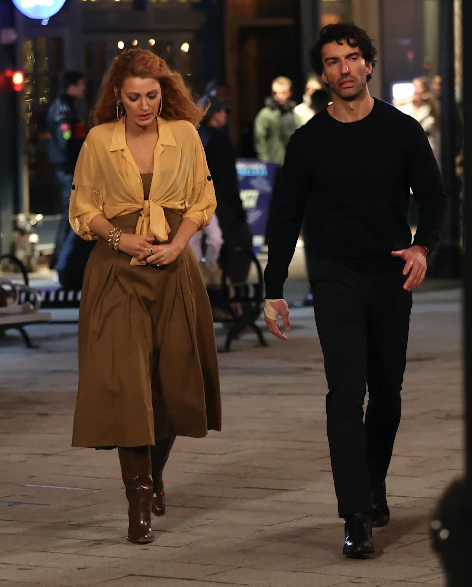 A woman in a stylish blouse and long skirt walks alongside a man in dark attire on a city street at night