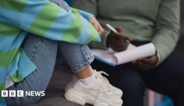 A person on the left is wearing jeans, a blue and green striped top, and white trainers. They are sitting on a chair with their legs pulled up. To their right is a person with a green top and navy trousers sitting in a chair with a clipboard and a pen. Neither person's head and shoulders is visible.