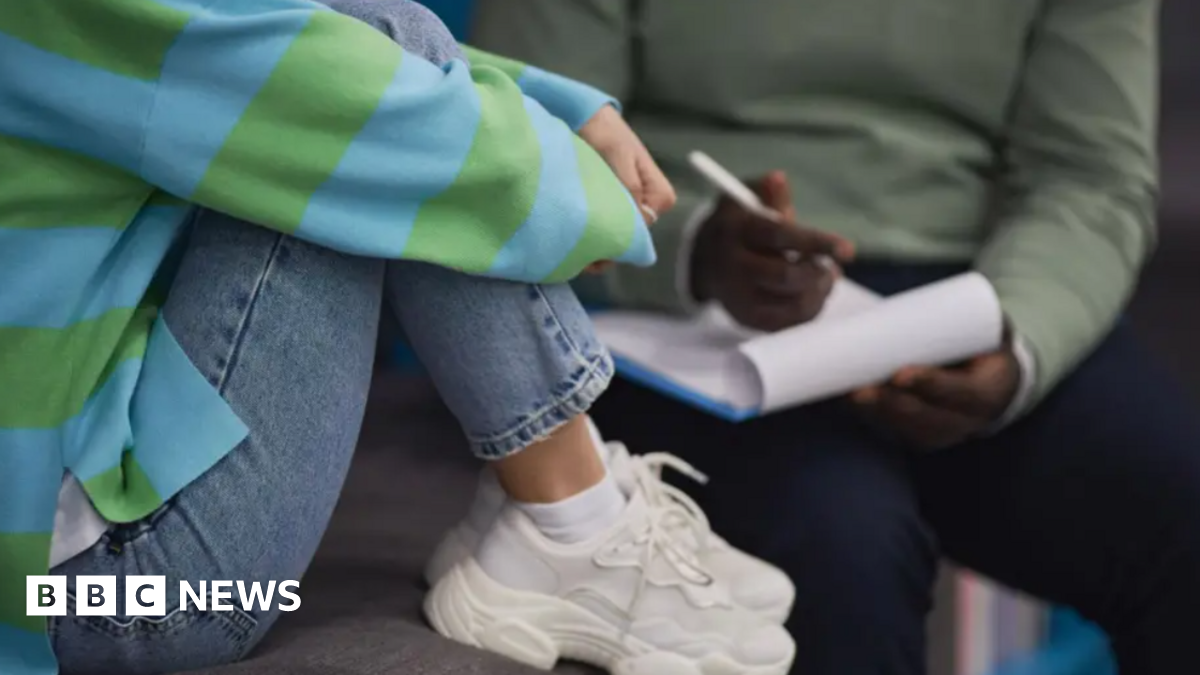 A person on the left is wearing jeans, a blue and green striped top, and white trainers. They are sitting on a chair with their legs pulled up. To their right is a person with a green top and navy trousers sitting in a chair with a clipboard and a pen. Neither person's head and shoulders is visible.