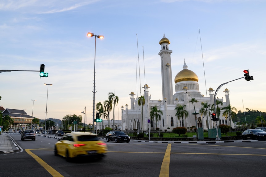 A white mosque with golden domed roofs seen alongside a road travelled by various cars.