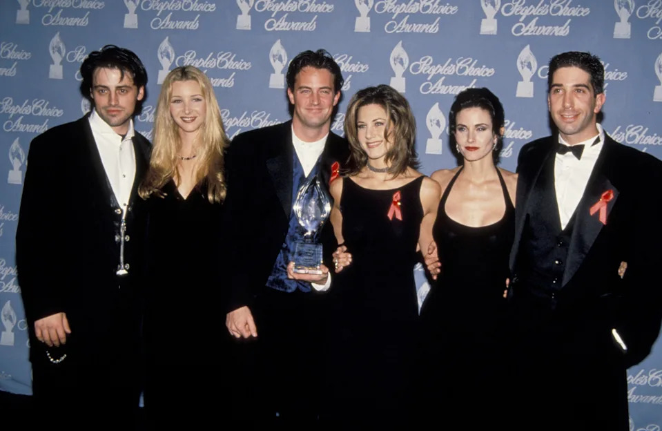 Group of actors in formal attire at awards event, holding a trophy, standing against a backdrop with "People's Choice Awards" text