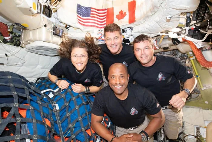 Four astronauts in a spacecraft pose for a photo, smiling. Two are standing and two are crouched in front, with American and Canadian flags visible
