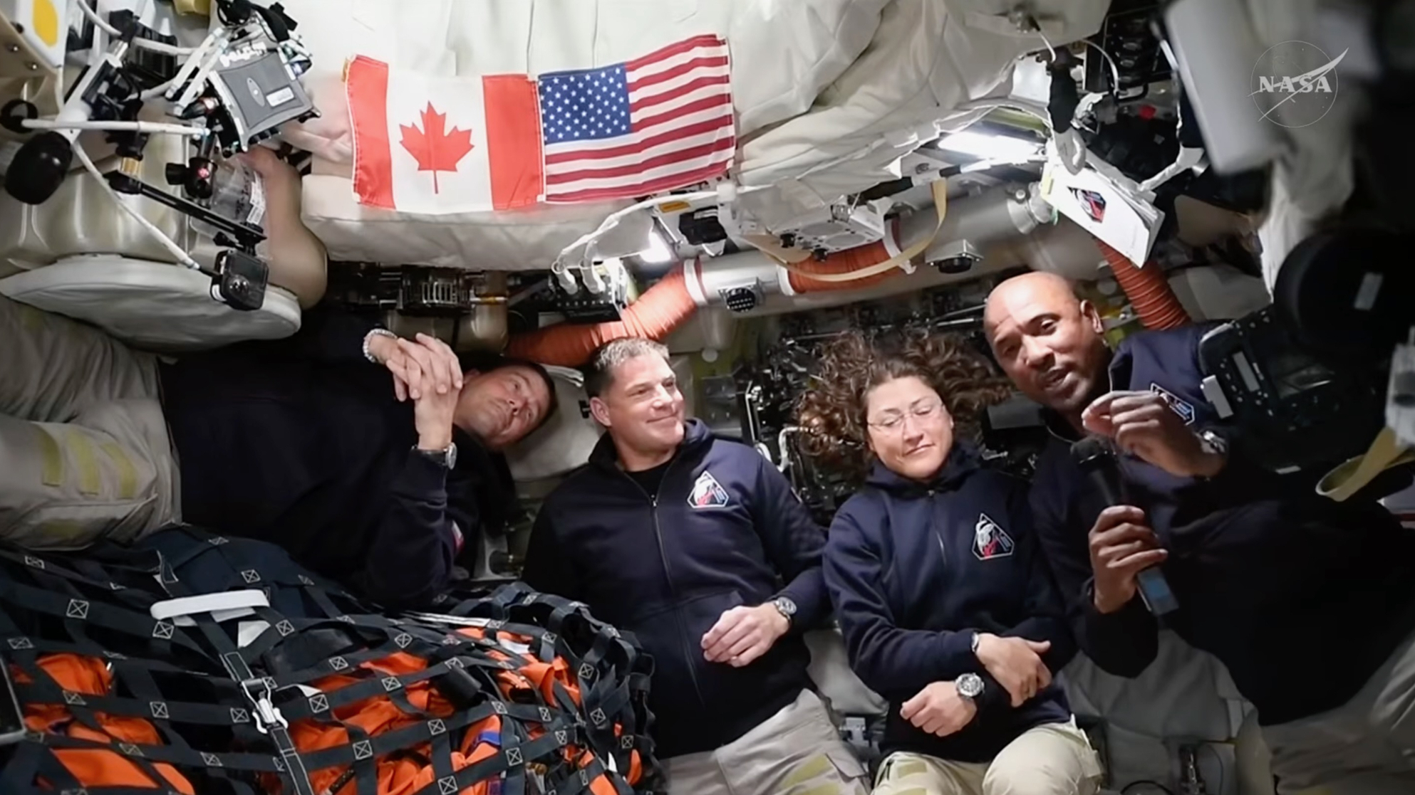 three male and one female astronaut, all wearing blue jackets and khaki pants, smile for the camera inside their space capsule in space