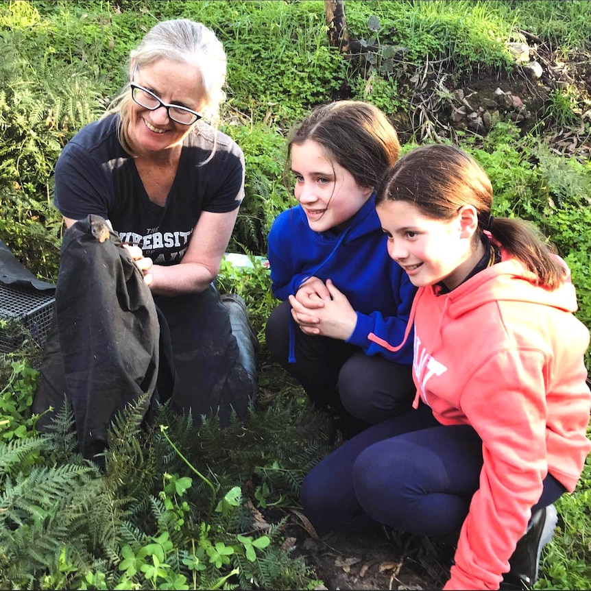 A woman and two young girls crouch on the ground with a blanket and small animal