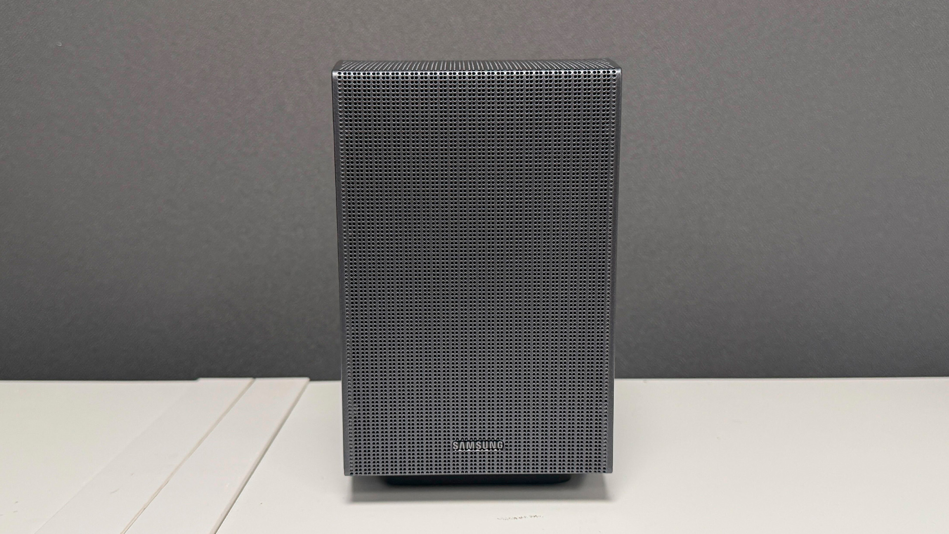 Samsung HW-Q990H soundbar package one surround speaker on white wooden surface in front of grey baffle