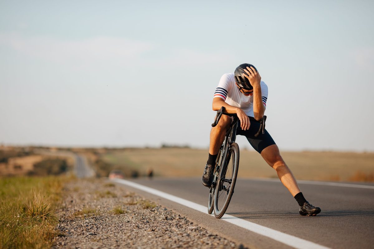 Exhausted cyclist in black helmet and sport clothing relaxing on road after long riding. Background of countryside. Concept of recreation.
