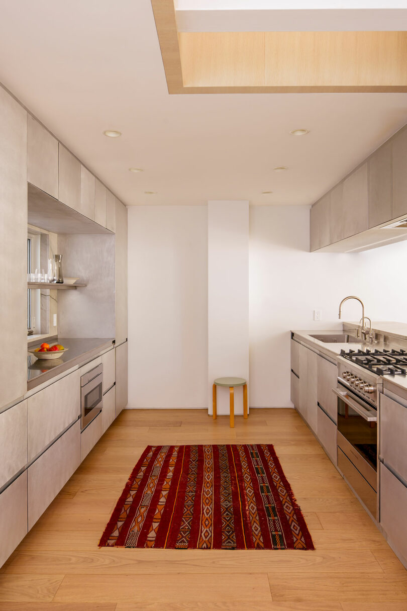 Modern galley kitchen with light wood floors, gray cabinetry, stainless steel appliances, and a red patterned rug in the center. A small stool sits against the far wall.