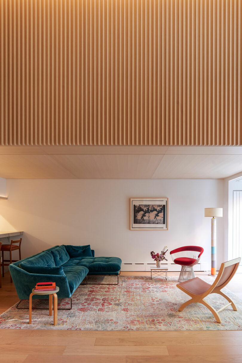 A modern living room with a teal sectional sofa, red chair, light wood furniture, patterned rug, standing lamp, and framed artwork on a cream wall with vertical wooden paneling above.
