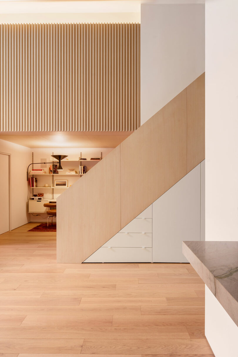 Minimalist interior with light wood flooring, a wooden staircase with built-in storage, and a small office nook with shelves and a desk beneath the upper level.