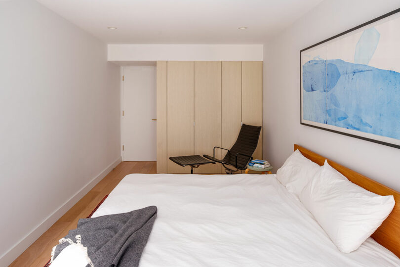 Minimalist bedroom with a made bed, black chair with books, light wood closet, white walls, and a large abstract blue artwork above the bed.