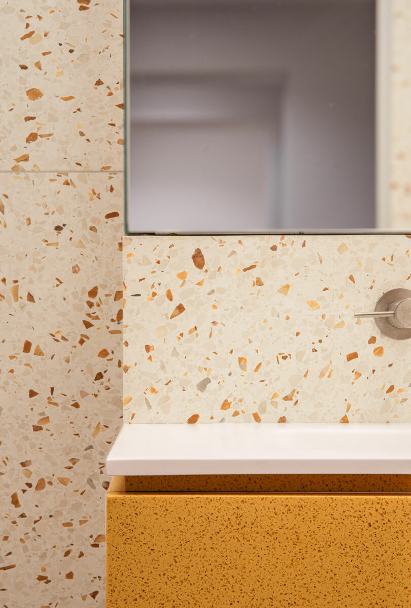 Close-up of a bathroom vanity with a terrazzo wall, a rectangular mirror, part of a white sink, a round metal fixture, and a yellow cabinet drawer.
