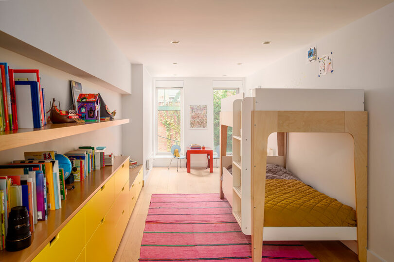 A brightly lit children's bedroom with a bunk bed, yellow storage drawers, bookshelves, a pink striped rug, and a small table near large windows.