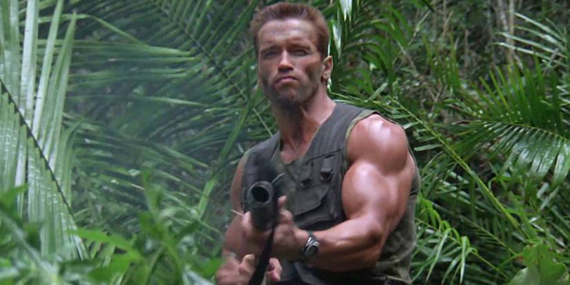 Arnold Schwarzenegger as Dutch in Predator aiming a machine gun while standing in front of jungle foliage
