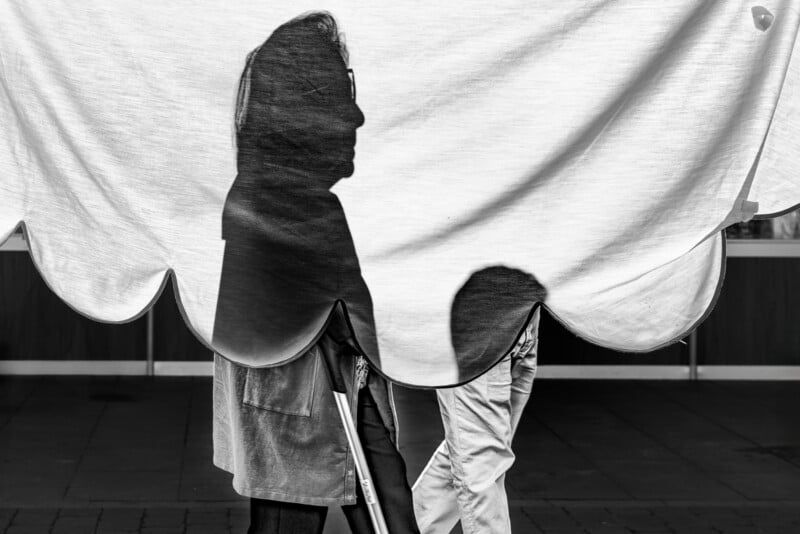 A black and white photo shows the silhouette of a person's head and upper body cast on a scalloped sheet, with another person walking behind the sheet, only their legs visible beneath it.