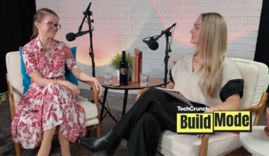 Two women sit facing each other in a podcast studio, speaking into microphones. One wears a red floral dress and glasses, while the other wears a cream top and black pants. A small table between them holds a bottle of wine, glasses of water, and books. A TechCrunch Build Mode logo appears in the corner.