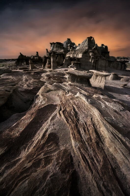 Eroded rock formations under a dramatic, orange-lit sky at dusk, with textured stone layers in the foreground and jagged hoodoos rising in the background.