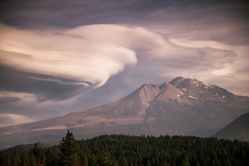 A forest stretches beneath a large mountain with patches of snow. Dramatic, swirling lenticular clouds fill the sky above the peak, illuminated with soft light, creating a surreal and serene landscape.