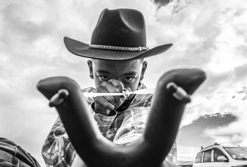 A boy in a cowboy hat aims a slingshot directly at the camera, with intense focus in his eyes. The photo is in black and white, and a car and cloudy sky are visible in the background.
