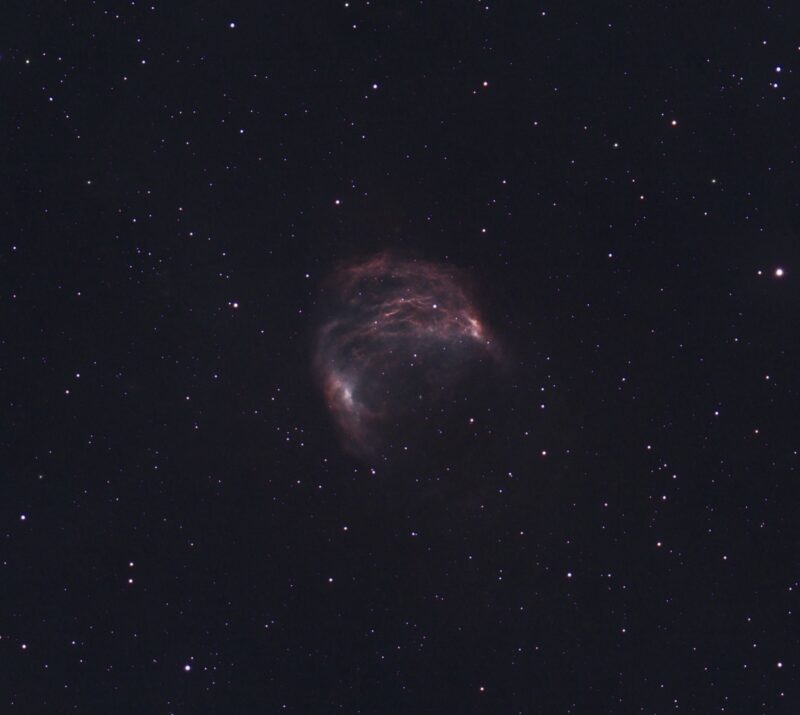 A broken cocoon of pink nebulosity with stars immersed within and in the surroundings.