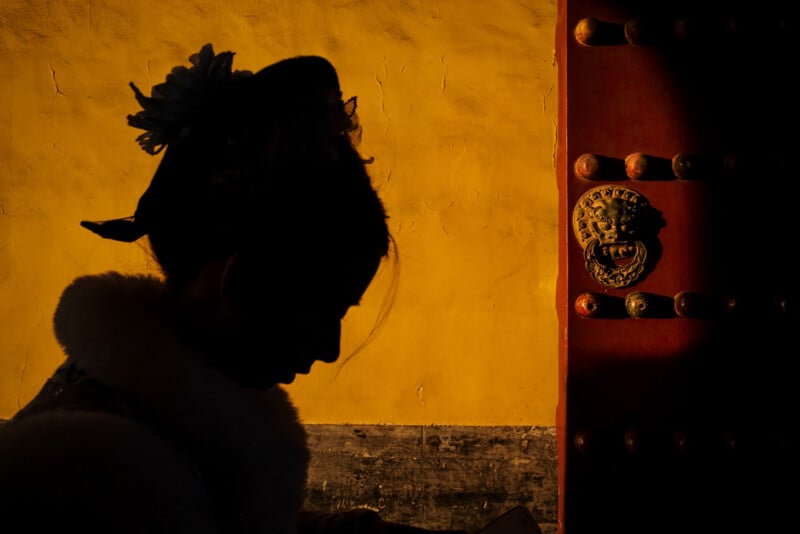 A silhouette of a person in traditional attire with a hair ornament stands near a red door with a lion head knocker, against a yellow wall illuminated by sunlight.