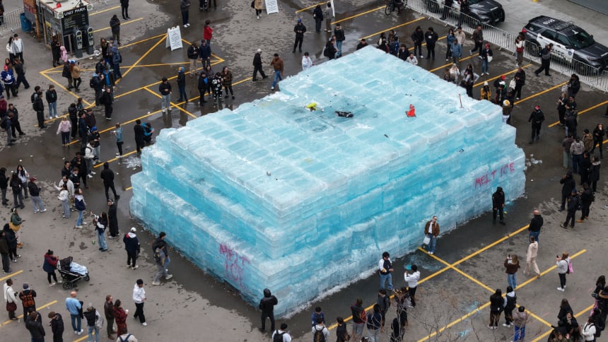 A large ice block stands in the middle of a parking lot, with people gathered around it.