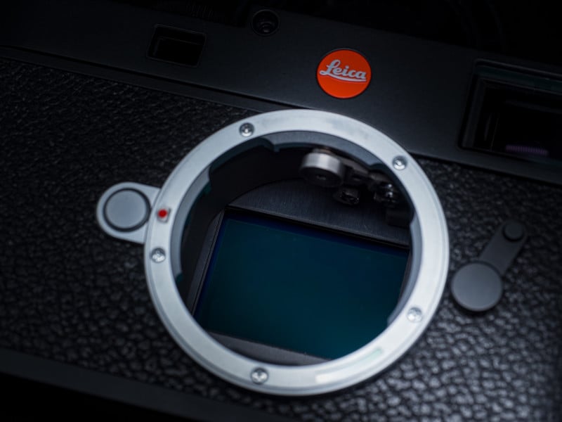 Close-up of a Leica camera body without a lens, showing the lens mount and sensor. The red Leica logo is visible at the top, and the camera has a textured black surface.