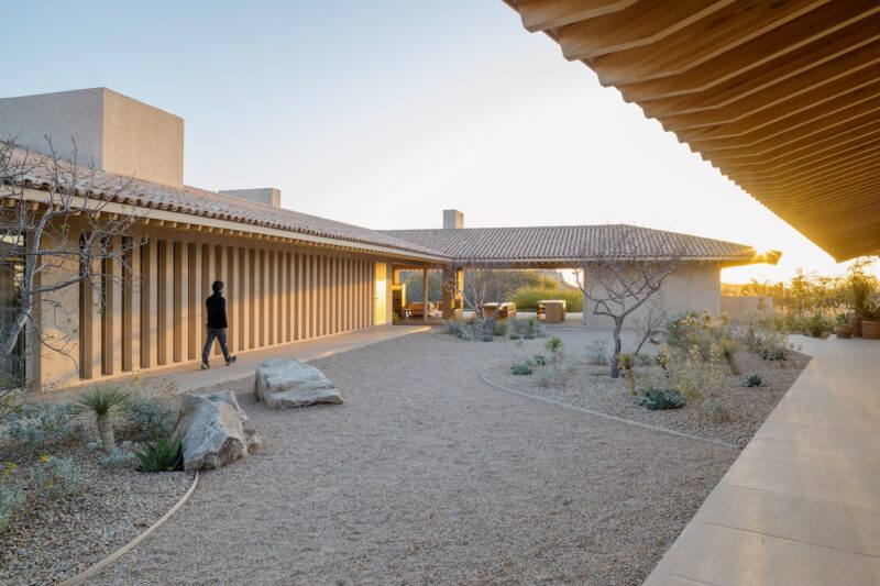 A person walks through a modern courtyard with gravel, rocks, and desert plants, surrounded by buildings with tiled roofs at sunset.