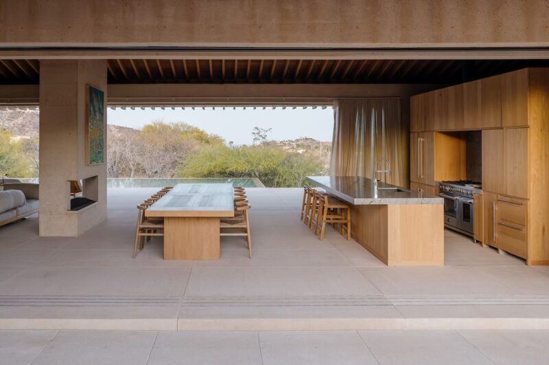 Modern open-plan kitchen and dining area with wood cabinetry and furniture, large windows, and views of trees and hills in the background.