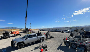 A wide view of an enhanced geothermal systems site. Various workers, trucks, trailers, and equipment are visible against a blue sky.