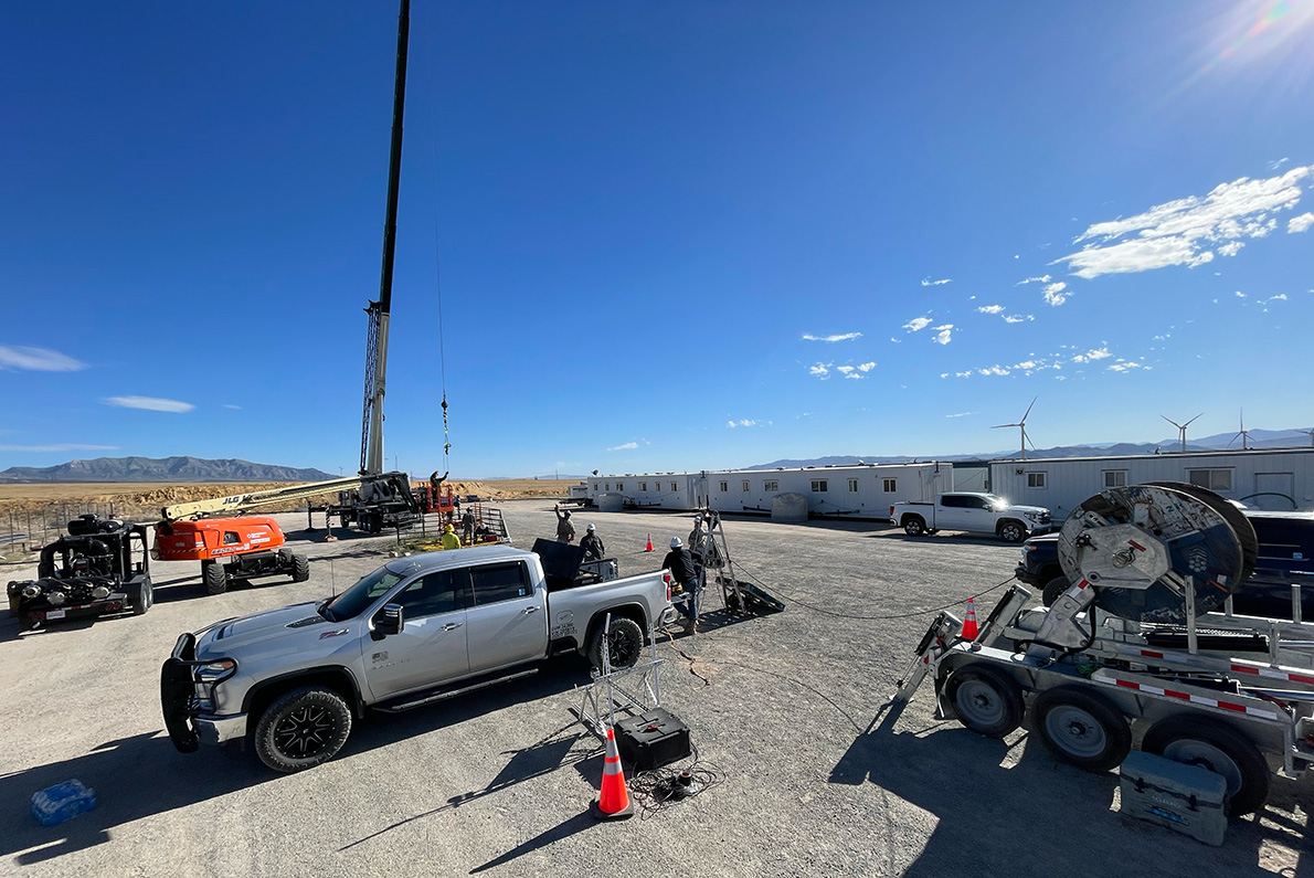 A wide view of an enhanced geothermal systems site. Various workers, trucks, trailers, and equipment are visible against a blue sky.