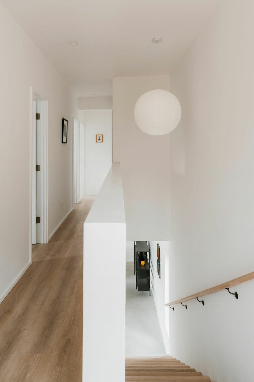 A minimal, modern hallway with light wood flooring, white walls, a round ceiling light, doors on the left, and stairs with a wooden handrail on the right.