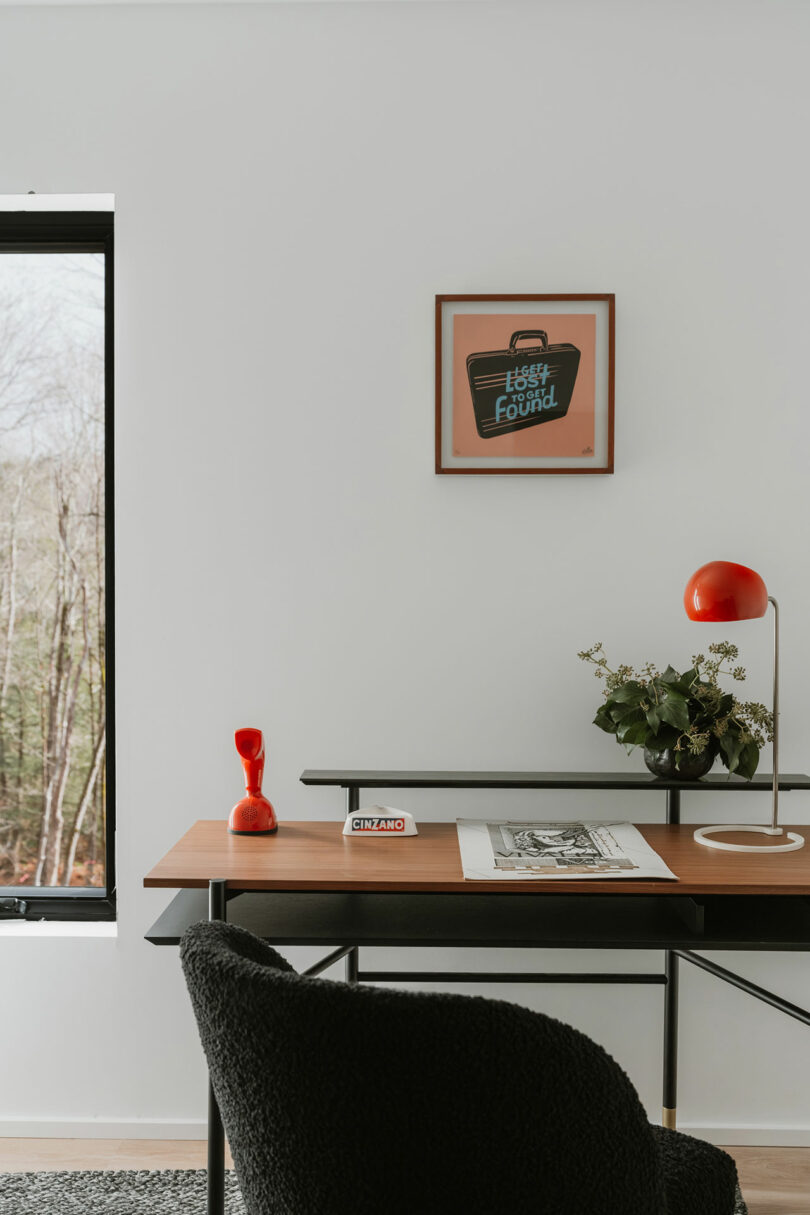 A modern desk with a black chair, red lamp, plant, and framed art on a white wall near a window overlooking trees.