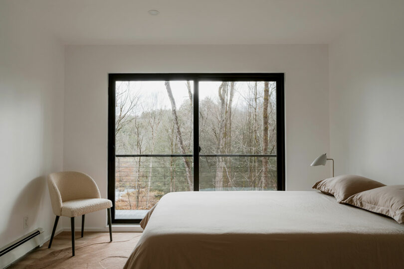 Minimalist bedroom with a beige chair, bed, lamp, and large window overlooking trees and a balcony.
