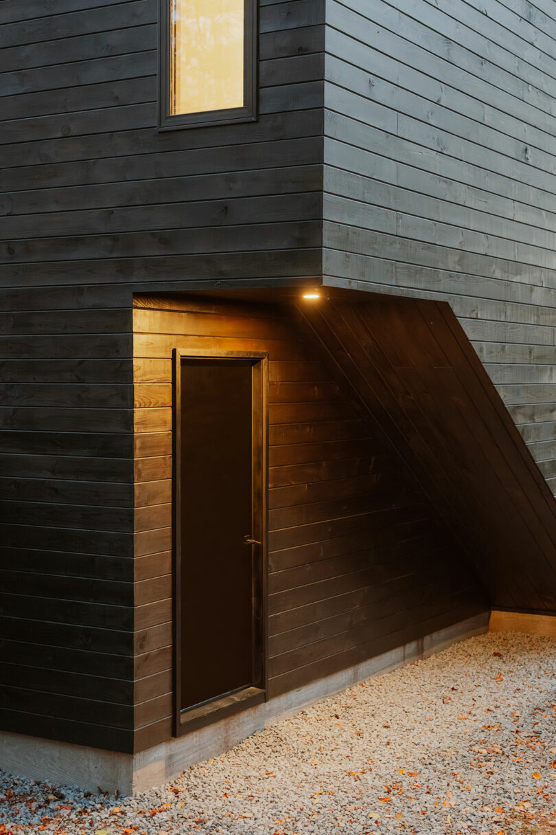 A modern black wooden exterior features a closed door under an exterior light, with a gravel ground and a single window above.