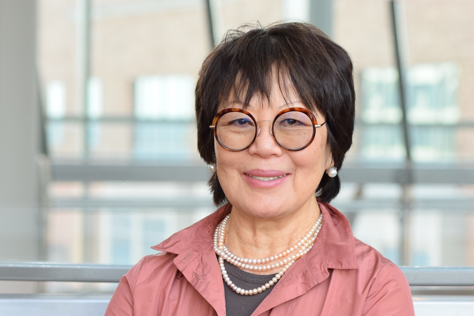A woman with short brown hair wearing a pink jacket, round glasses and pearls smiles at the camera.