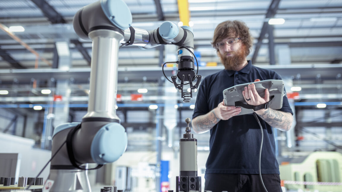 Image of a man with a computer tablet standing in front of an industrial robotic arm.