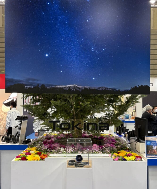 A display booth featuring a Tokina camera lens, surrounded by colorful flowers and greenery, with a large starry night sky photo and mountain landscape above the booth. People are visible in the background.