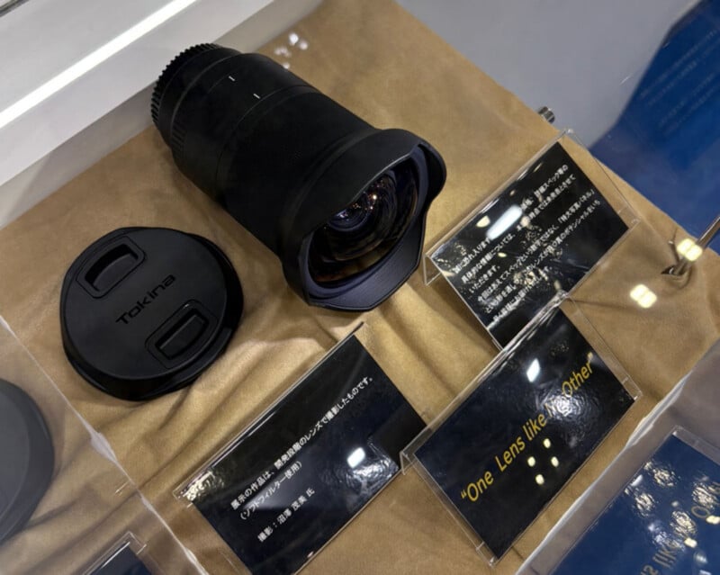 A wide-angle Tokina camera lens and its lens cap are displayed on a brown surface, alongside informational plaques, one reading "One Lens like no other!" in yellow text.