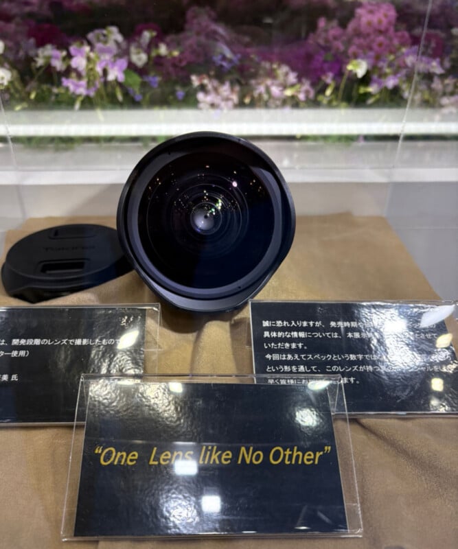 A camera lens is displayed upright on a brown surface, flanked by informational placards in Japanese. The front placard reads: “One Lens like No Other” in yellow text. Purple flowers are visible in the background.