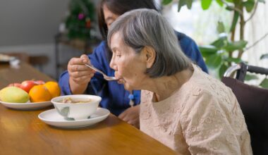 Caregiver feeding elderly woman with soup in dining room.