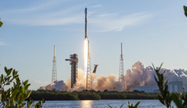 Image shows a rocket launching vertically in a morning blue sky with white clouds at Cape Canaveral Space Force Station in Florida on Saturday, April 11, 2026. The launch reflects in a nearby body of water and shrubs. Photo credit: SpaceX