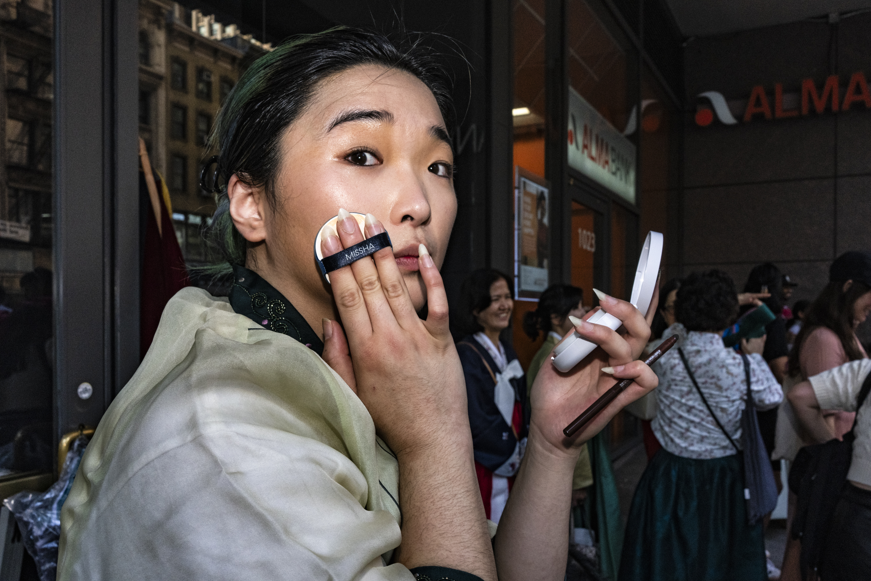 A young person with dark green-tinted hair applies foundation with a Missha cushion compact while holding a small mirror, standing on a busy urban street.