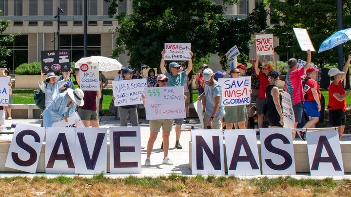 People hold signs outside on a sunny day in protest of NASA budget cuts, July 20, 2025.