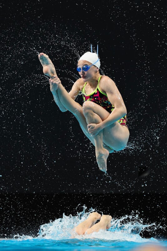 A synchronized swimmer in a colorful swimsuit and white cap performs a complex jump above a pool, tucking her legs while water splashes below and another swimmer's legs are visible at the surface.