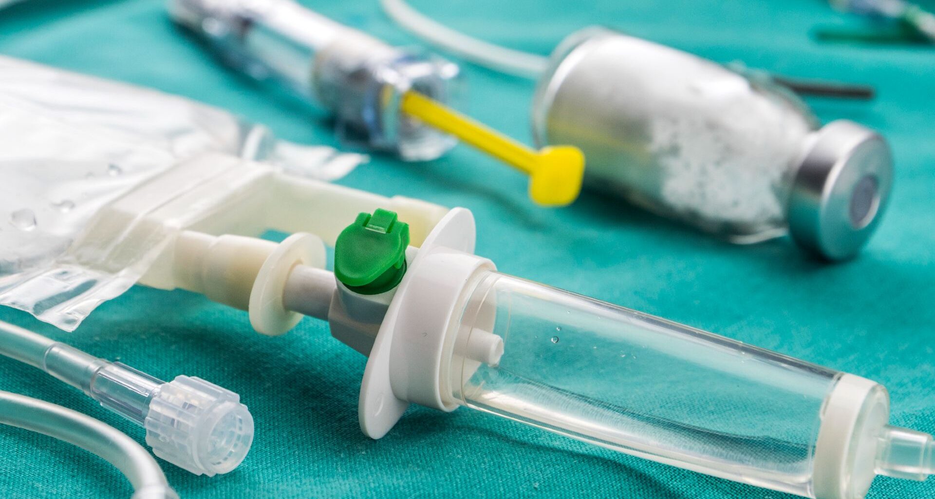 A close up of a series of medicine bottles, IV drips and syringes sitting on a turquoise sheet.